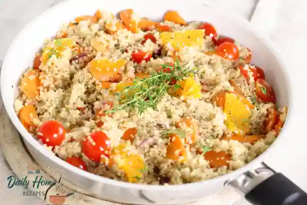 Colorful quinoa salad with cherry tomatoes, carrots, orange slices, and fresh thyme in a white pan.