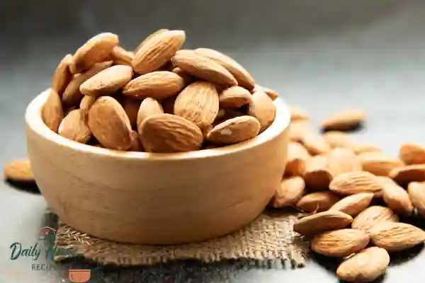 Raw whole almonds in a wooden bowl, with scattered almonds around on a dark surface.
