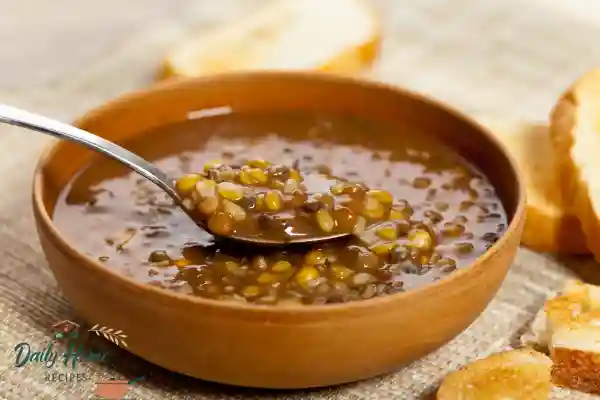 A wooden bowl of warm lentil soup with a spoon lifting a mix of cooked green and brown lentils, served with sliced bread.